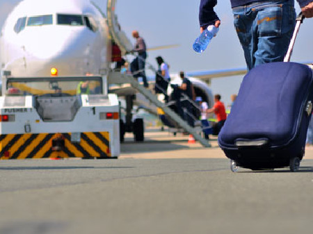 Mann zieht einen blauen Rollkoffer zu einem Flugzeug, bei dem mehrere Passagiere &uuml;ber die Treppe einsteigen