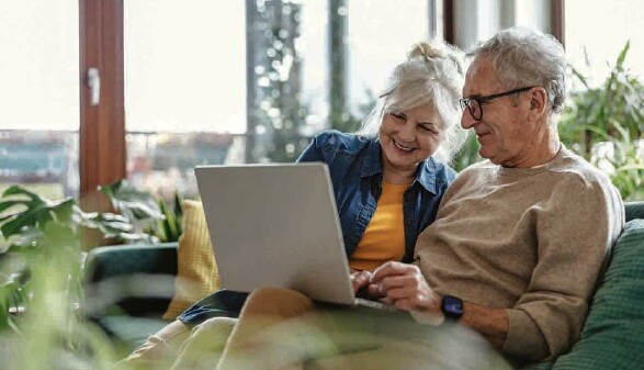 Älteres Ehepaar sitzt lächelnd auf einer Couch mit einem geöffneten Laptop auf den Knien