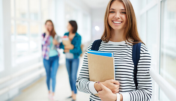 M&auml;dchen h&auml;lt eine Mappe in H&auml;nden und steht auf einem Schulgang. Hinter ihm sieht man unscharf zwei junge Frauen, die ebenfalls Sch&uuml;lerinnen sein d&uuml;rften.
