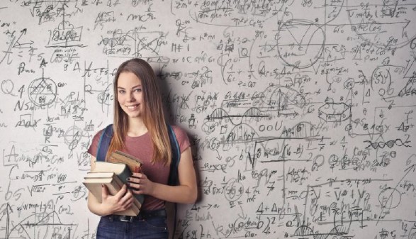 Eine junge Frau steht mit einem Stapel Bücher am Arm an einer Wand, die mit Formeln vollgekritzelt ist. © Canva