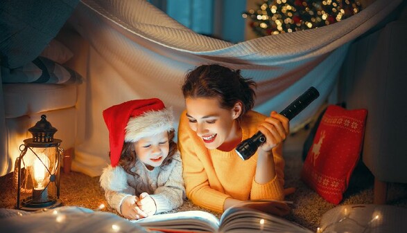 Mutter und Kind mit Weihnachtsmütze sitzen mit einer Taschenlampe vor einem Buch