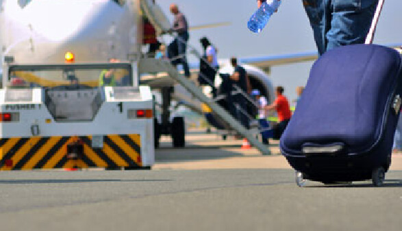 Mann zieht einen blauen Rollkoffer zu einem Flugzeug, bei dem mehrere Passagiere &uuml;ber die Treppe einsteigen