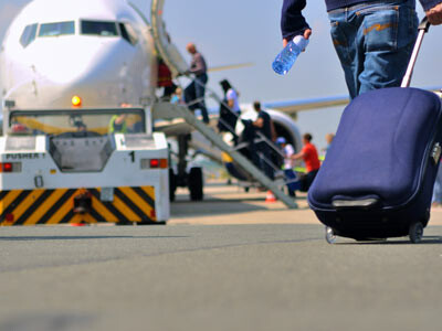 Mann zieht einen blauen Rollkoffer zu einem Flugzeug, bei dem mehrere Passagiere &uuml;ber die Treppe einsteigen