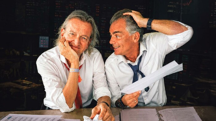 Markus Linder und Hubert Trenkwalder pr&auml;sentieren verzweifeln bei der Schreibtischarbeit, im Hintergrund sind Kurse an der B&ouml;rse zu sehen