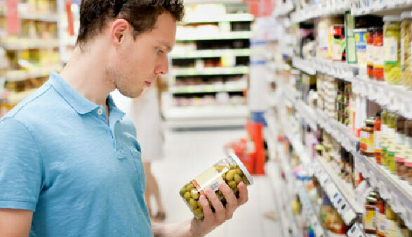 Ein junger Mann steht vor einem Supermarkt-Regal und schaut sich das Etikett auf einem Glas mit grünen Oliven an.