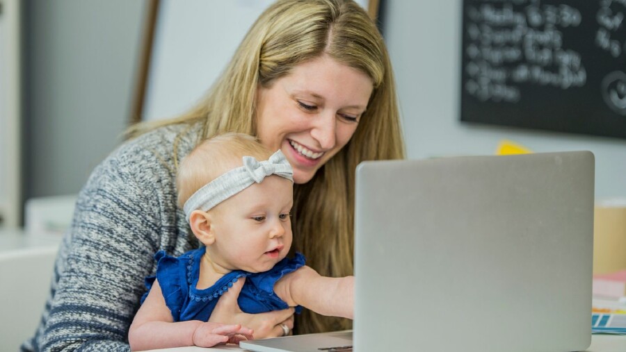 Eine blonde Frau sitzt mit Baby am Schoß vor dem Laptop und lacht, weil die Kleine tippen möchte.