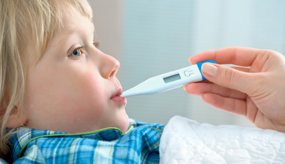Ein kleines Kind liegt im Bett, man sieht die Hand einer Erwachsenen, welche dem Kind ein Fieberthermometer in den Mund steckt.