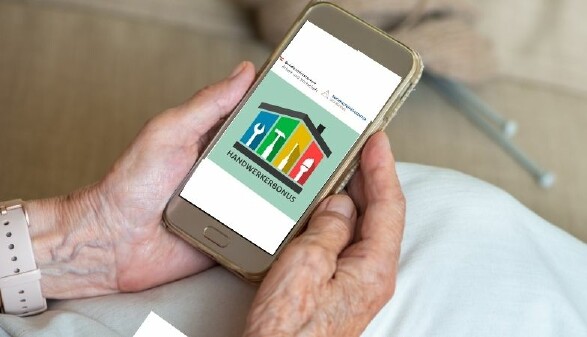 Ein Smartphone in den Händen einer älteren Person, darauf die Website zum Handwerkerbonus.