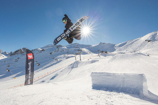 Snowboarder springt über die Schanze und posiert in der Luft, im Hintergrund Sonnenschein
