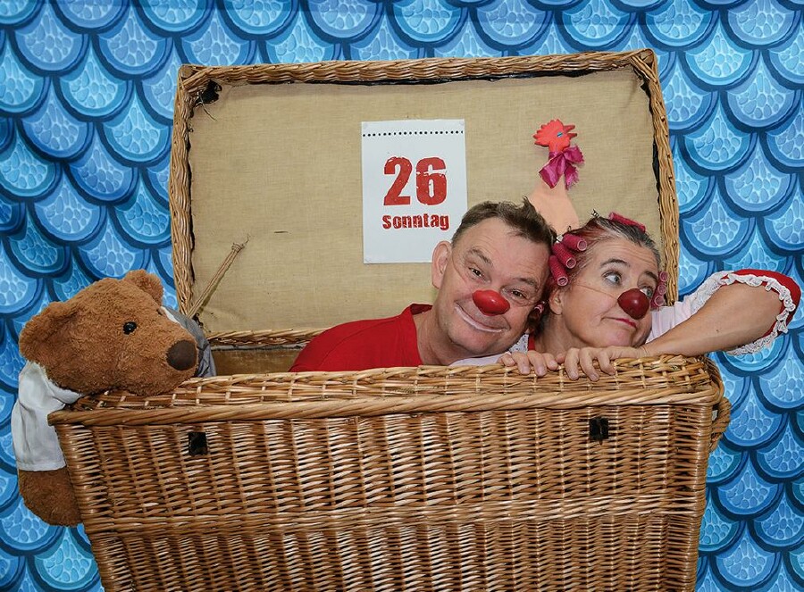 Die Clowns Herbert und Mimi sitzen mit roten Nasen in einem geflochtenen Reisekoffer. Links ist ein brauner Stoffbär. Im Hintergrund ist ein blau gemustertes Tuch gespannt.