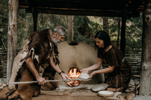 Ein Paar ist mit Tierfellen bekleidet und bereitet einen Brotlaib zum Backen vor. Im Hintergrund lodert Feuer in einem gemauerten Backofen