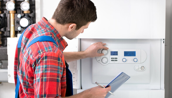 Ein Techniker mit rotkariertem Hemd und blauer Latzhose steht vor einer Heizungsanlage und bedient mit der linken Hand einen Regler-Knopf. In der rechten Hand h&auml;lt er einen Kugelschreiber und eine blaue Mappe.