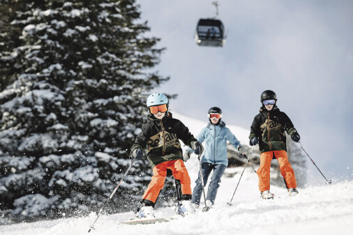 Drei Personen auf der Piste beim Skifahren, über ihnen fährt die Gondel auf den Berg 