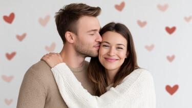 Vor einem Hintergrund mit roten und rosa Herzen steht ein j&uuml;ngeres Liebespaar. Links steht der Mann, er tr&auml;gt einen hellbraunen Pulli. Er hat kurzes hellbraunes Haar und einen leichten Bartansatz. Er dreht seinen Kopf nach rechts und k&uuml;sst die Frau, die neben ihm steht, mit geschlossenen Augen auf die Schl&auml;fe. Die Frau ist etwas kleiner, tr&auml;gt einen wei&szlig;en Pulli, hat langes braunes Haar. Sie legt ihre linke Hand auf die rechte Schulter des Mannes und l&auml;chelt in die Kamera.