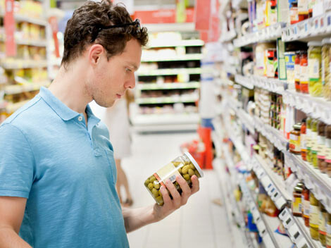 Ein junger Mann steht vor einem Supermarkt-Regal und schaut sich das Etikett auf einem Glas mit grünen Oliven an.