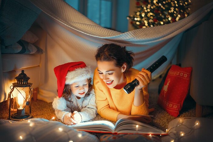 Mutter und Kind mit Weihnachtsmütze sitzen mit einer Taschenlampe vor einem Buch