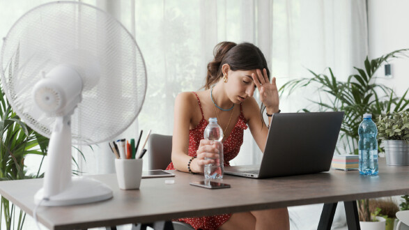 Junge Frau in leichtem Sommerkleid sitzt am Schreibtisch und wischt sich Schweiß von der Stirn. Vor ihr ein Ventilator und zwei Wasserflaschen.