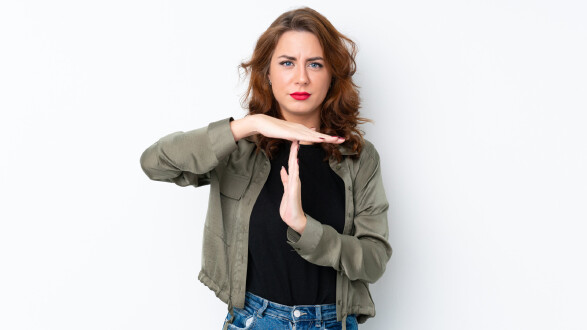 Frau mit welligem braunen Haar und ernstem Blick steht vor einer weißen Wand und macht mit ihren Händen das Zeichen für Auszeit.