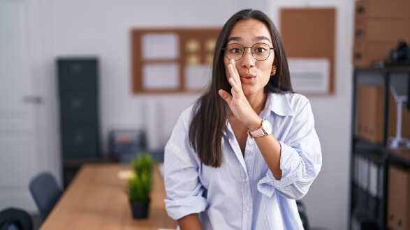Junge Frau in einem Büro erzählt mit vorgehaltener Hand ein Geheimnis