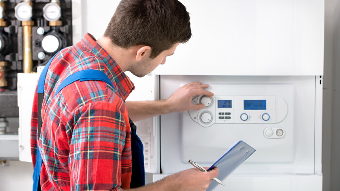 Ein Techniker mit rotkariertem Hemd und blauer Latzhose steht vor einer Heizungsanlage und bedient mit der linken Hand einen Regler-Knopf. In der rechten Hand h&auml;lt er einen Kugelschreiber und eine blaue Mappe.