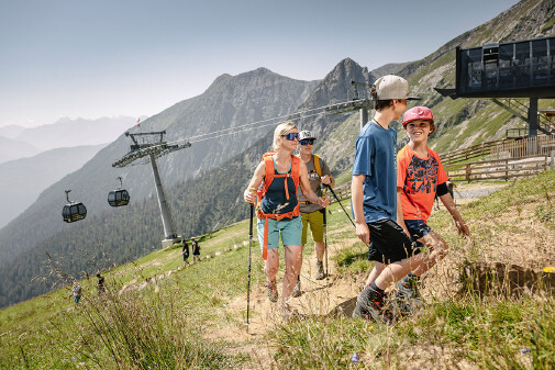 Eine vierk&ouml;pfige Familie wandert neben der Seilbahn-Trasse bergauf, im Hintergrund sind Wanderer bergab zu sehen