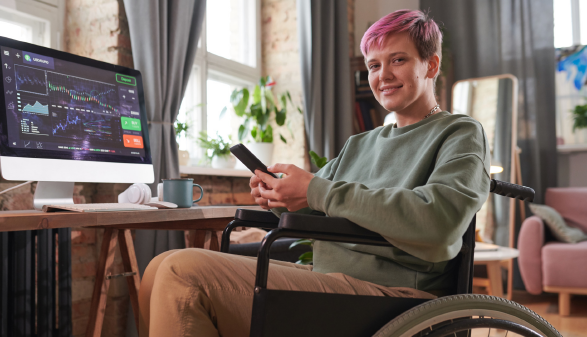 Junge Frau mit kurzen pinken Haaren sitzt mit Handy in der Hand im Rollstuhl vorm Computer, auf dem eine Seite mit Aktienkursen geöffnet ist. Sie lächelt in die Kamera.