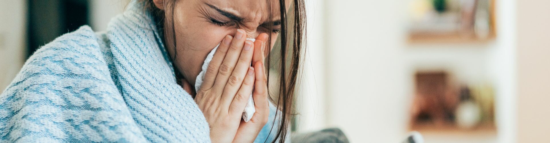 Portraitbild einer Dame, eingewickelt in eine hellblaue Decke, die sich gerade schnäuzt. 
