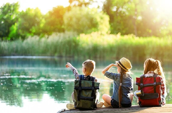 © New Africa/stock.adobe.com Ein Bub und zwei Mädchen sitzen am Ufer eines Sees