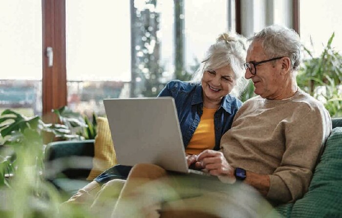 Älteres Ehepaar sitzt lächelnd auf einer Couch mit einem geöffneten Laptop auf den Knien