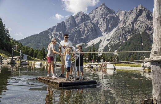 Eine vierk&ouml;pfige Familie steht auf einem kleinen Flo&szlig; und hantelt sich an einem Seil durch einen Teich