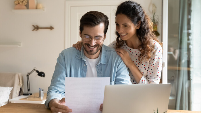 Mann sitzt am Schreibtisch und schaut freudig auf ein Blatt Papier, Frau umarmt ihn von hinten, liest mit und l&auml;chelt