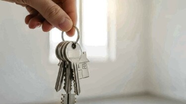 Eine linke Hand h&auml;lt einen Schl&uuml;sselbund, im Hintergrund sieht man einen leeren Raum und ein Fenster, durch das Licht einf&auml;llt.