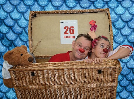 Die Clowns Herbert und Mimi sitzen mit roten Nasen in einem geflochtenen Reisekoffer. Links ist ein brauner Stoffbär. Im Hintergrund ist ein blau gemustertes Tuch gespannt.